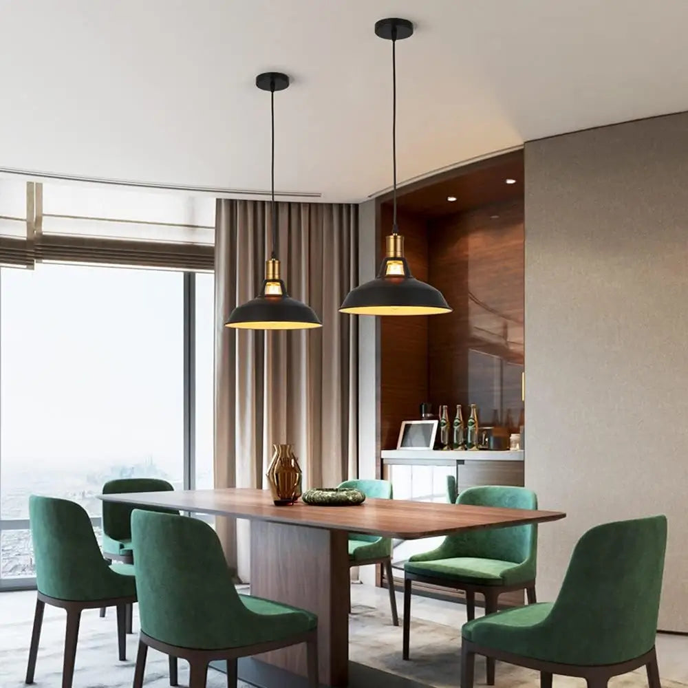 Two black industrial pendant lights with white interiors and brass accents hanging over a wooden dining table with green chairs in a modern dining room.