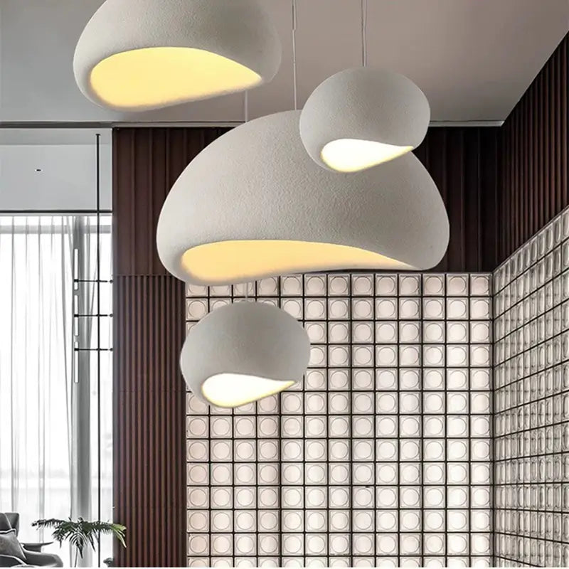 A cluster of assorted-sized modern minimalist pendant lights, featuring a textured, organic design, emitting warm ambient light in a contemporary interior with a unique glass block wall and wood paneling.
