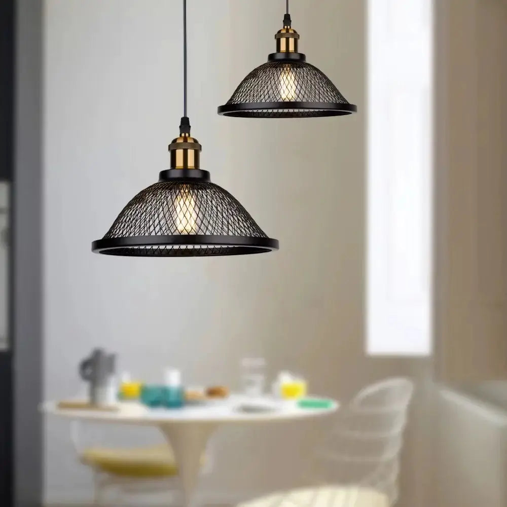 wo farmhouse industrial style pendant lights with black mesh domes and brass fittings, suspended above a white round dining table in a bright kitchen or dining area.