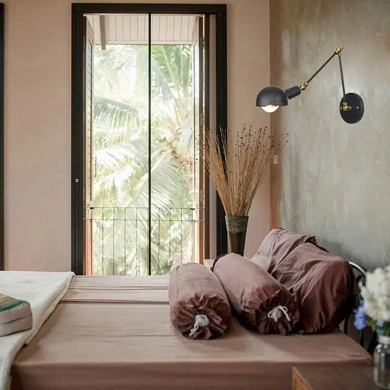 Industrial wall sconce with a black metal body and brass accents, mounted beside a cozy bedroom setting with soft lighting.