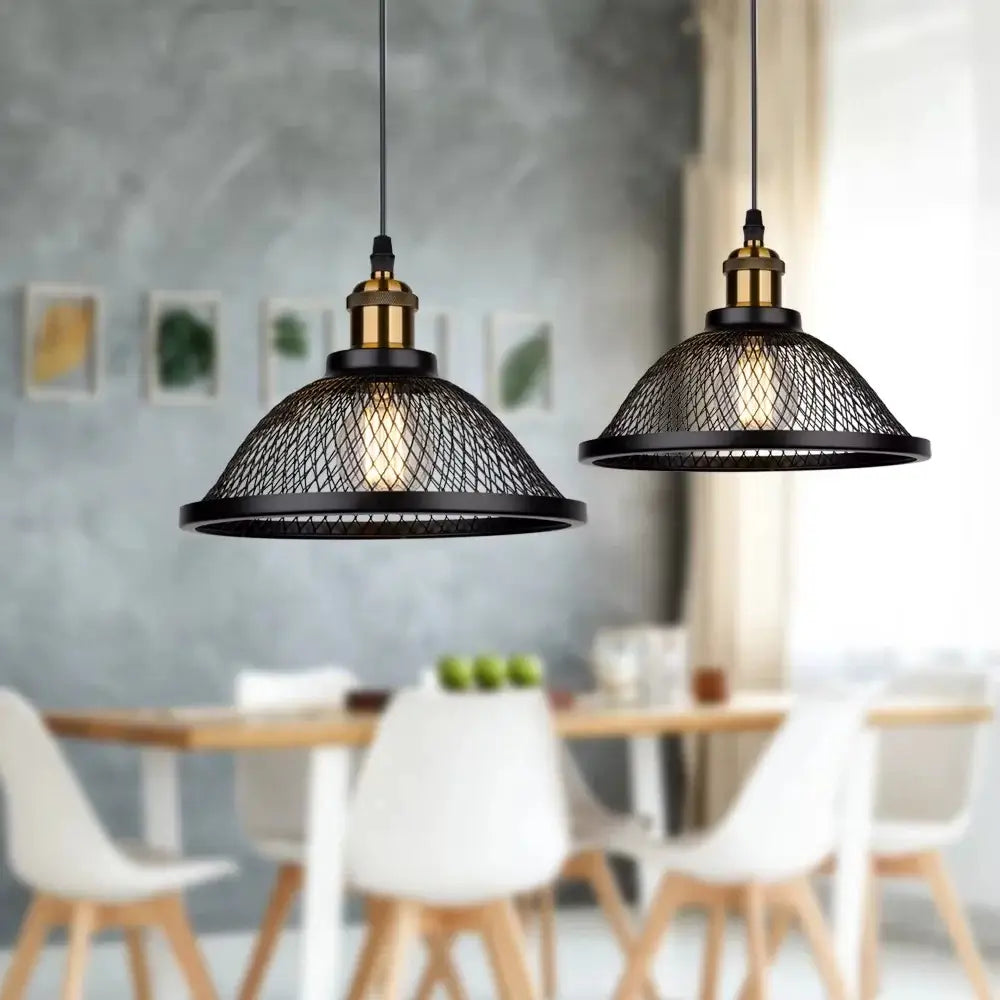Pair of modern industrial pendant lights with black mesh shades and brass accents, illuminating a contemporary dining room table with chairs in the background.