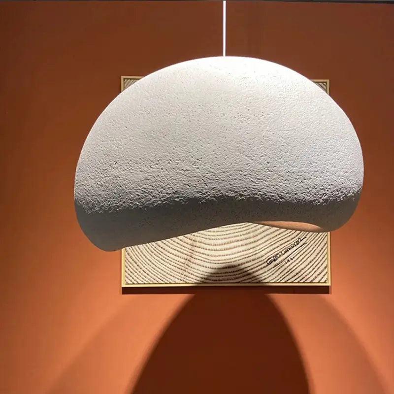A modern minimalist textured pendant light with an organic, asymmetric design, casting a warm glow and shadow against an orange wall, highlighting its sculptural form.