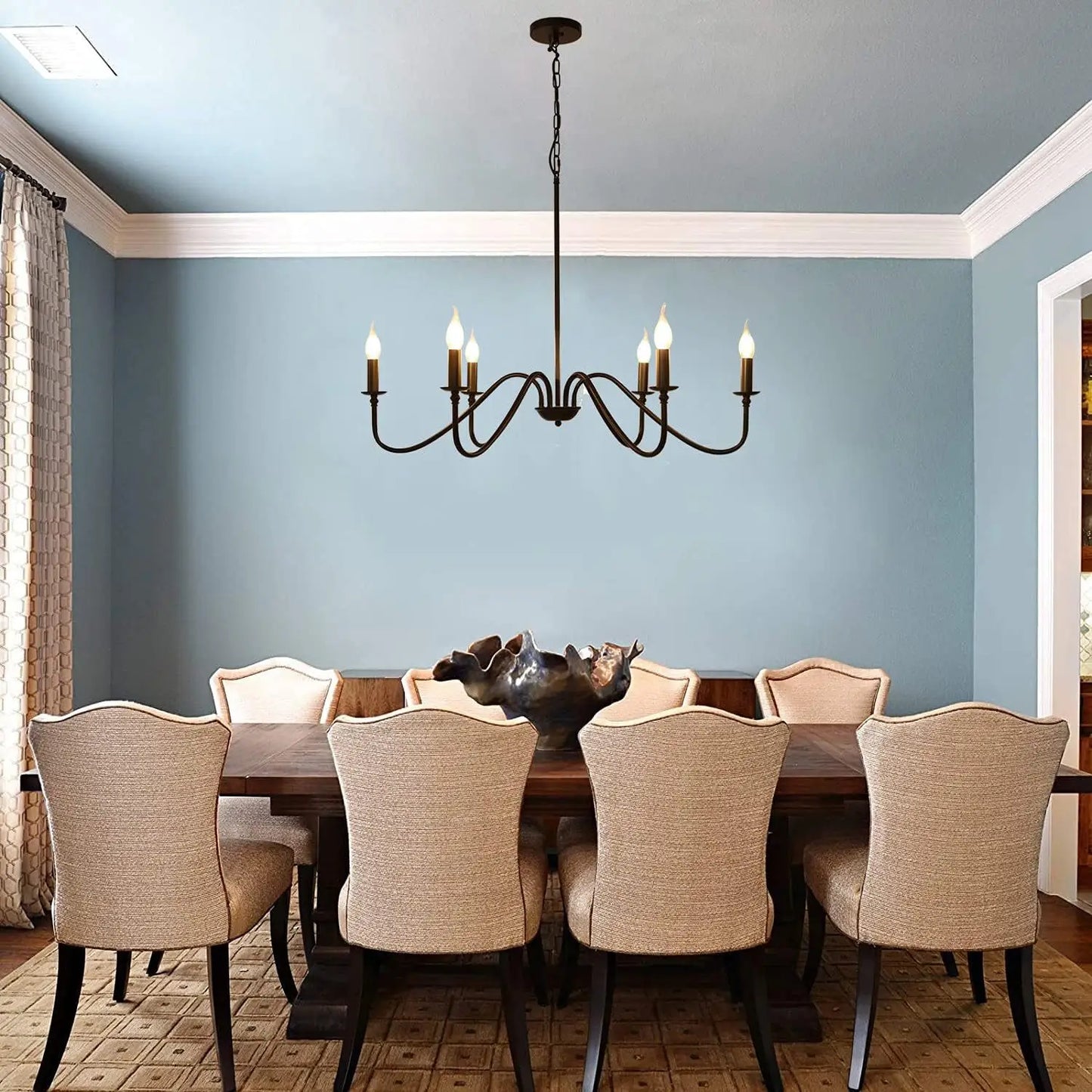 A rustic black 6-light chandelier in a dining room with a distinctive blue ceiling and a large wooden table.