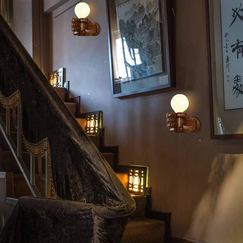 Industrial wall lamp in a modern design featuring a hand sculpture holding a glowing bulb, ideal for stairways or living room decor.