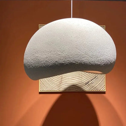 A modern minimalist textured pendant light with an organic, asymmetric design, casting a warm glow and shadow against an orange wall, highlighting its sculptural form.
