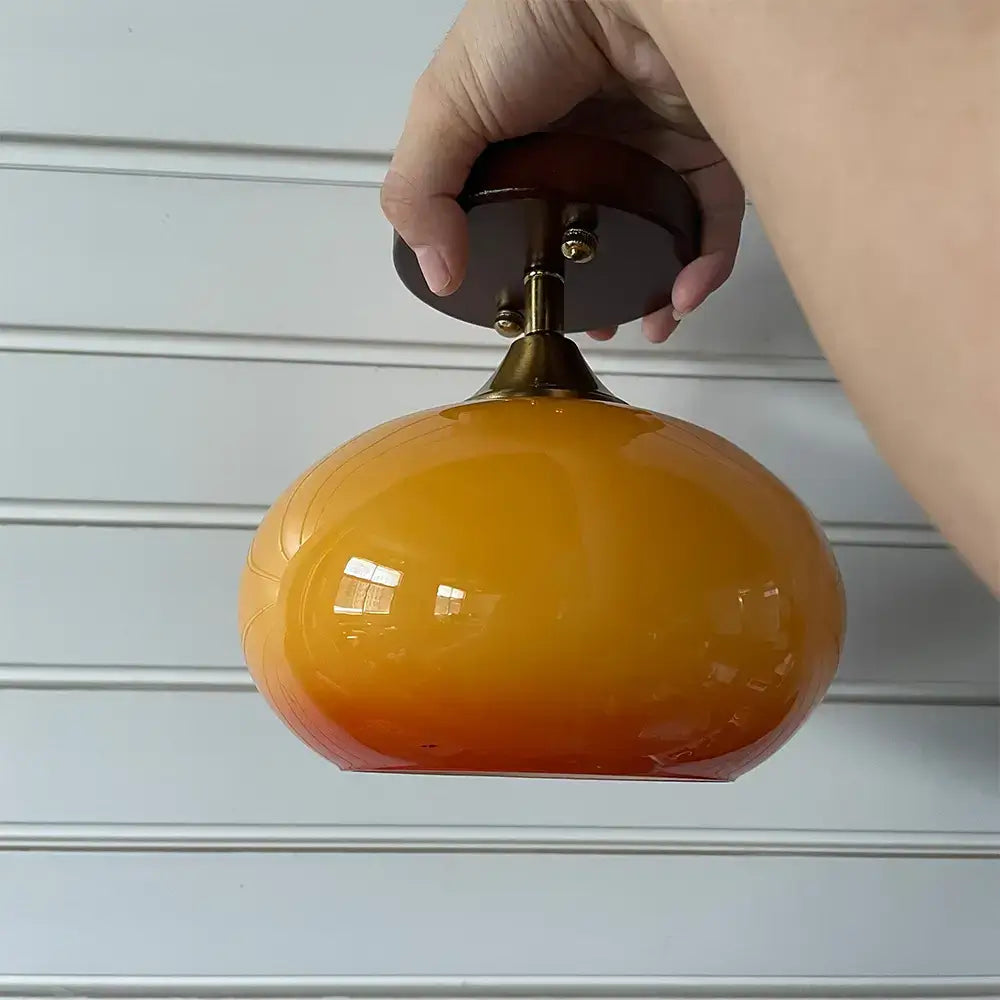 Hand holding a mid-century modern semi-flush mount ceiling light with a glossy orange glass shade and walnut wood canopy.